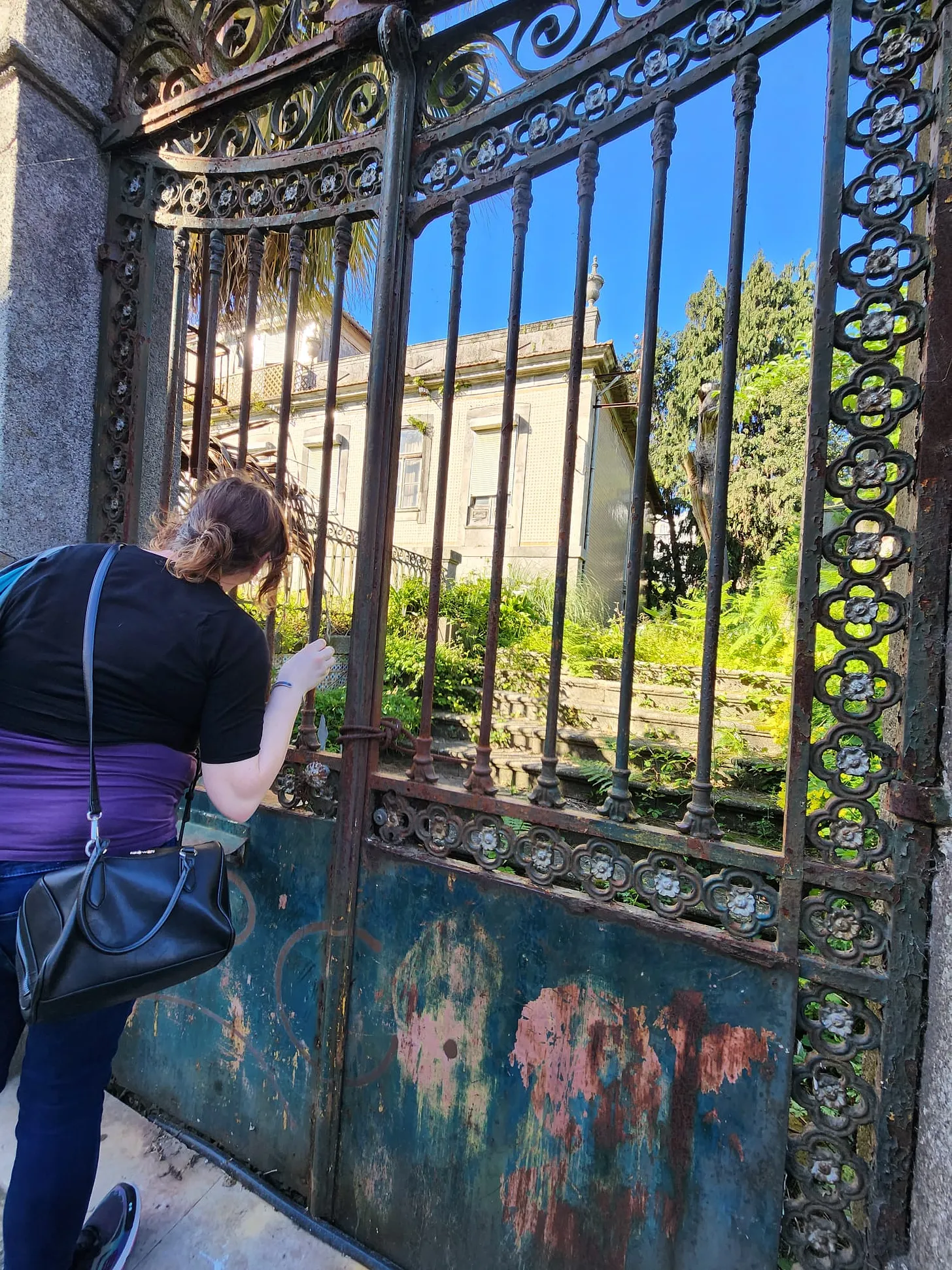 A woman peers through an ornate, rusty gate to a once stately estate, now abandoned, being reclaimed by nature