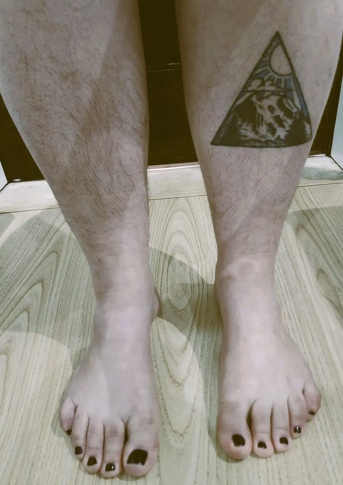two hairy legs with a tattoo of a mountain scene one one leg and toe nails painted dark red