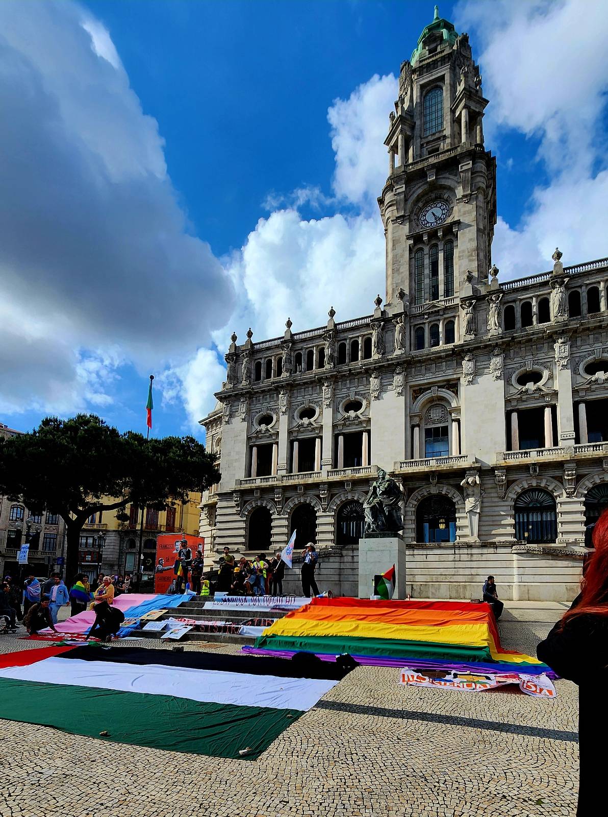 On the steps of city hall in Porto, Portugal, a large rainbow LGBTQ+ pride flag and a transgender pride flag are laid out next to a large Palestinian flag as protesters gather